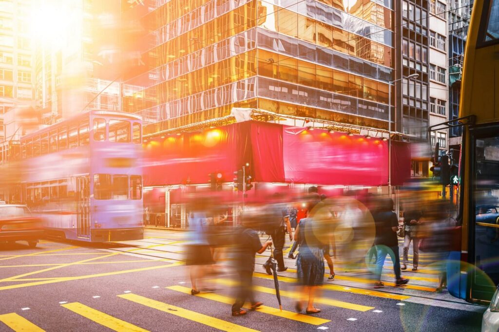 Blurred pedestrians crossing a busy Asian city street with a double-decker tram at sunset.